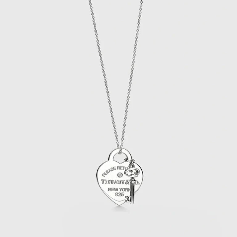 A silver heart-shaped pendant necklace with a key charm, featuring engraved text that reads 'PLEASE RETURN TO TIFFANY & CO. NEW YORK 925'.