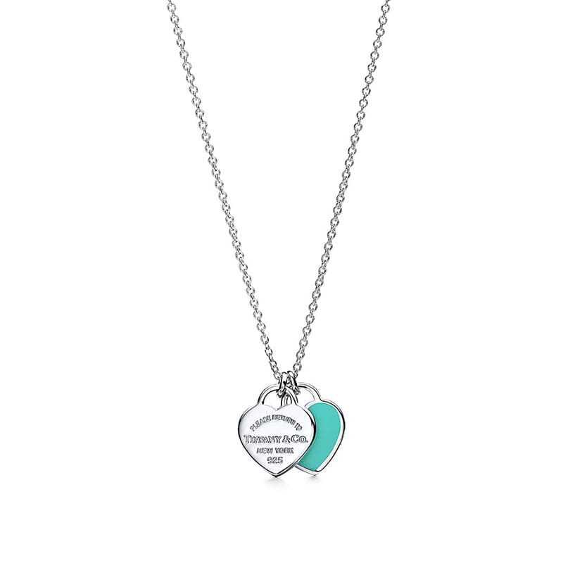 A silver chain necklace with two interlocking heart-shaped tags, one engraved with 'Tiffany & Co. NEW YORK 1904' and the other featuring a vibrant turquoise enamel inlay, shown on a plain white background.